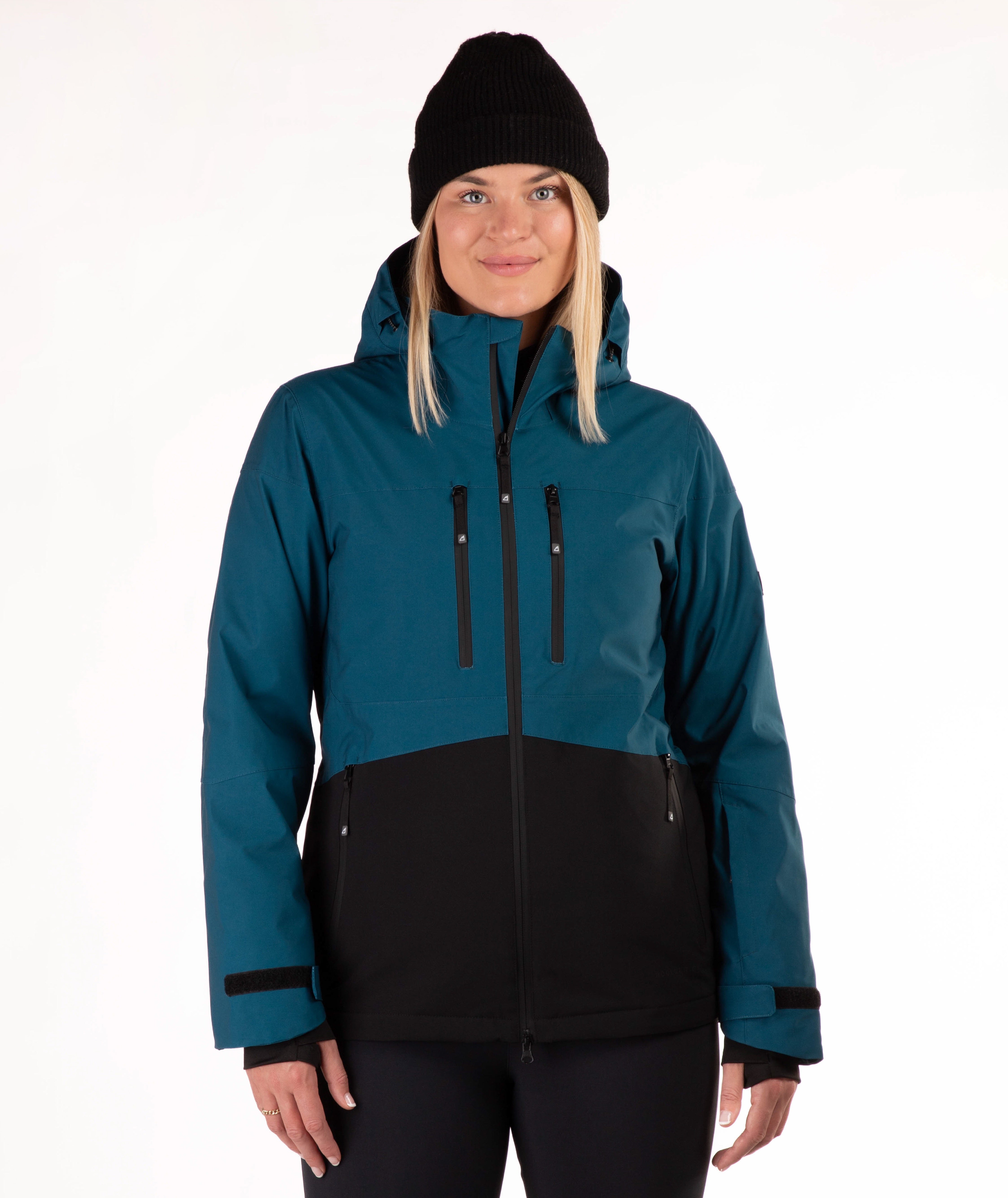 A woman wearing a dark teal and black color-blocked ski jacket, a black beanie, and black pants, standing against a plain white studio background.