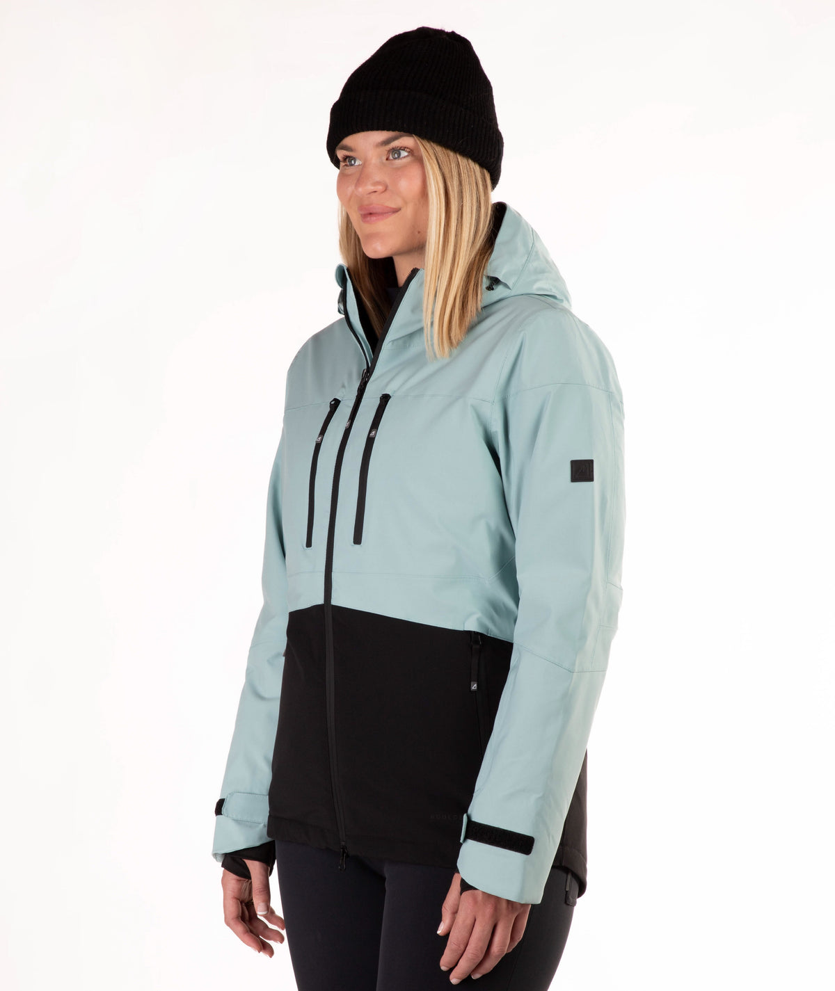 A woman wearing a light green and black ski jacket, a black beanie, and black pants, standing against a plain white studio background.