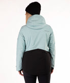 A woman wearing a light green and black ski jacket, a black beanie, and black pants, standing against a plain white studio background.