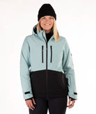 A woman wearing a light green and black ski jacket, a black beanie, and black pants, standing against a plain white studio background.