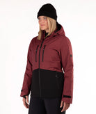 A woman wearing a color-blocked ski jacket in burgundy and black, along with a black beanie, photographed against a plain white studio background.