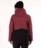 A woman wearing a color-blocked ski jacket in burgundy and black, along with a black beanie, photographed against a plain white studio background.