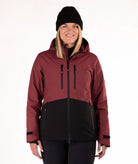 A woman wearing a color-blocked ski jacket in burgundy and black, along with a black beanie, photographed against a plain white studio background.