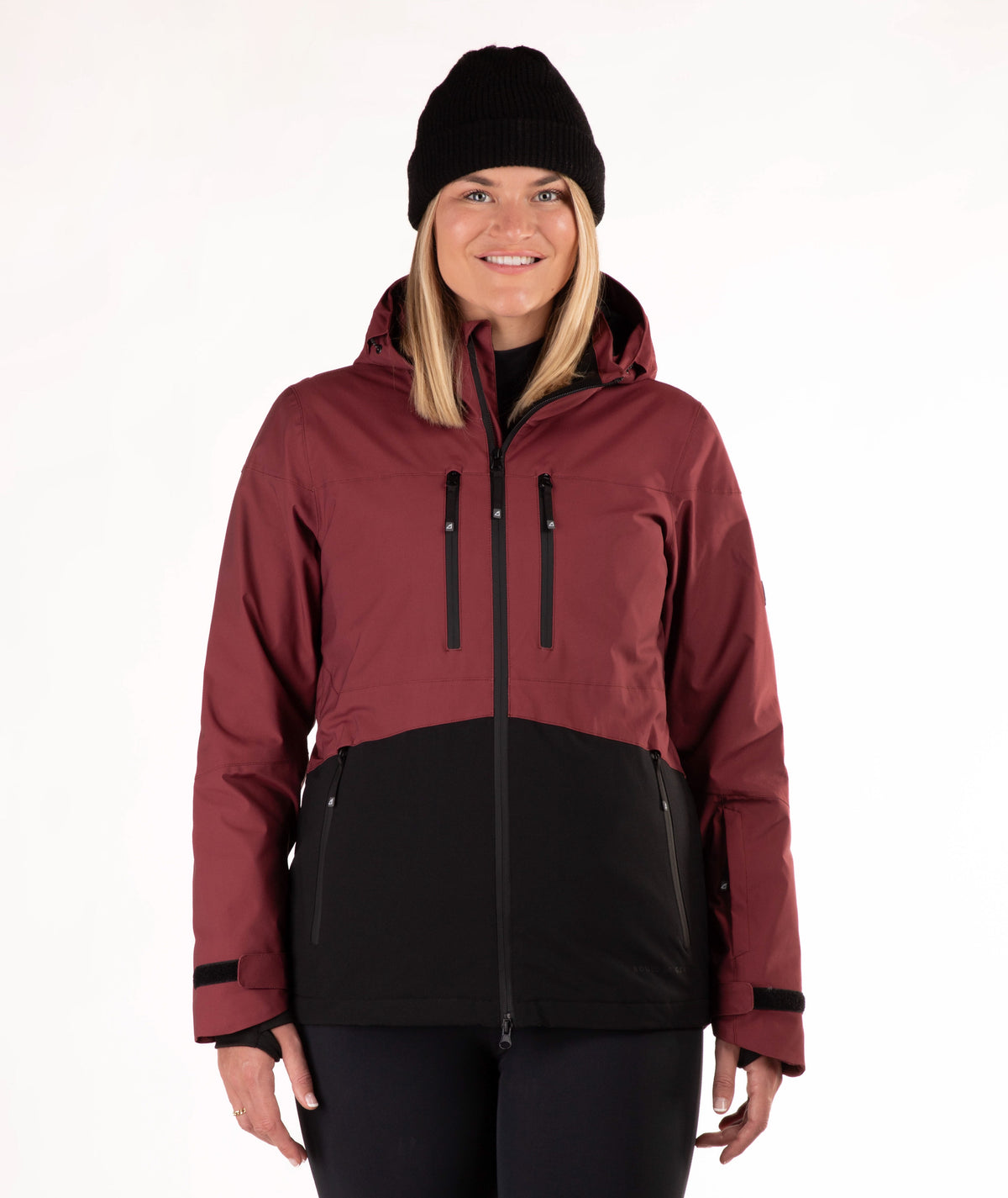 A woman wearing a color-blocked ski jacket in burgundy and black, along with a black beanie, photographed against a plain white studio background.
