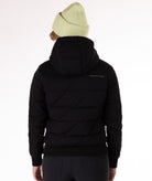 A woman wearing a black quilted jacket with a hood and black knit trim on wrist cuffs and hem on a white background. Back view of the jacket showing Boulder Gear brand logo.