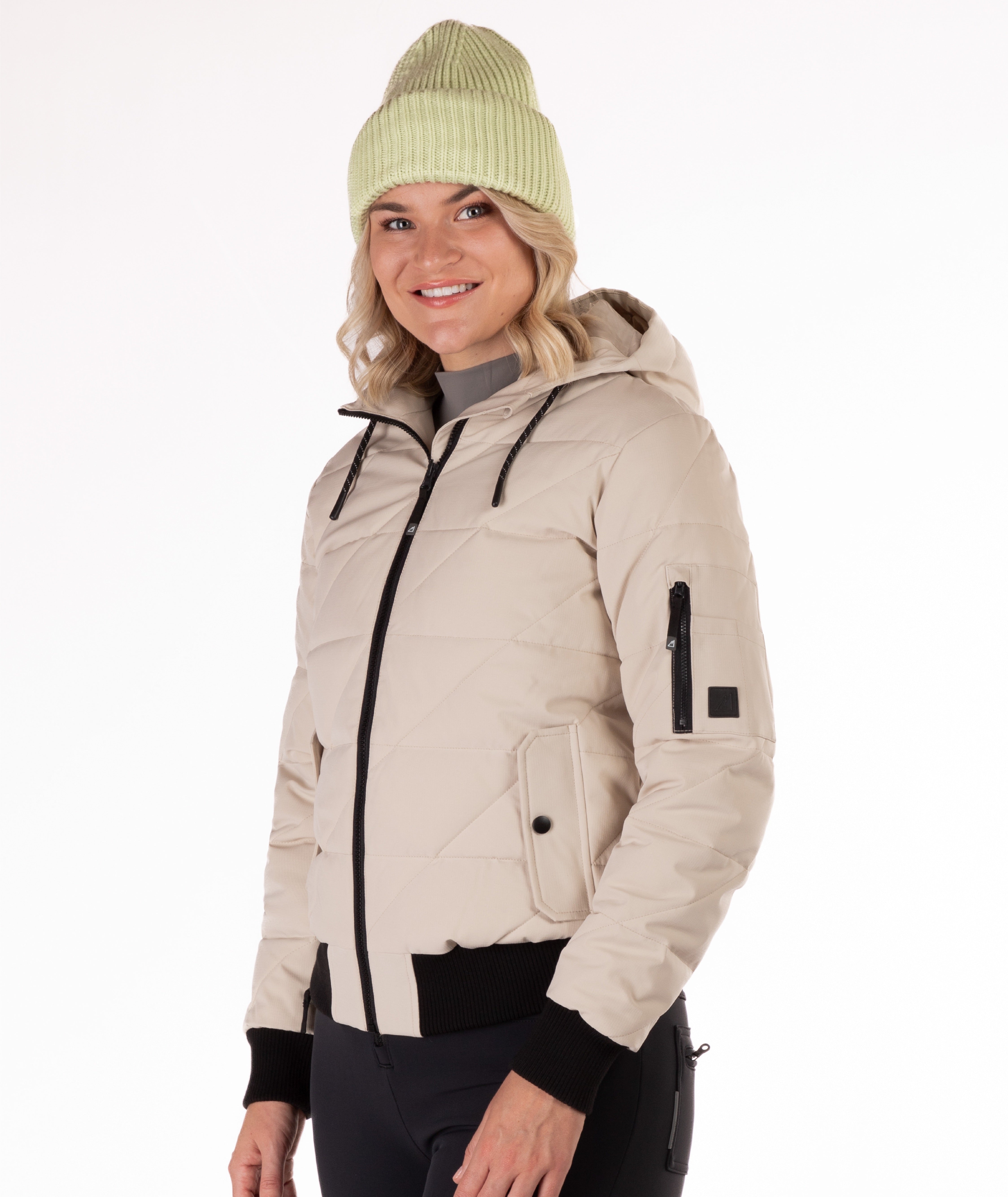 Woman wearing a beige quilted jacket with black knit trim on the sleeve cuffs and bottom hem on a white background. Side Profile view highlighting a cargo pocket on the left sleeve. 
