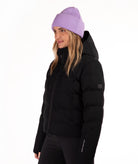 A woman wearing a black hooded puffer jacket and a light purple ribbed knit beanie, shown in profile against a plain white studio background.