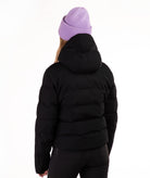 A person shown from the back wearing a black hooded puffer jacket, black pants, and a light purple ribbed knit beanie against a plain white studio background.