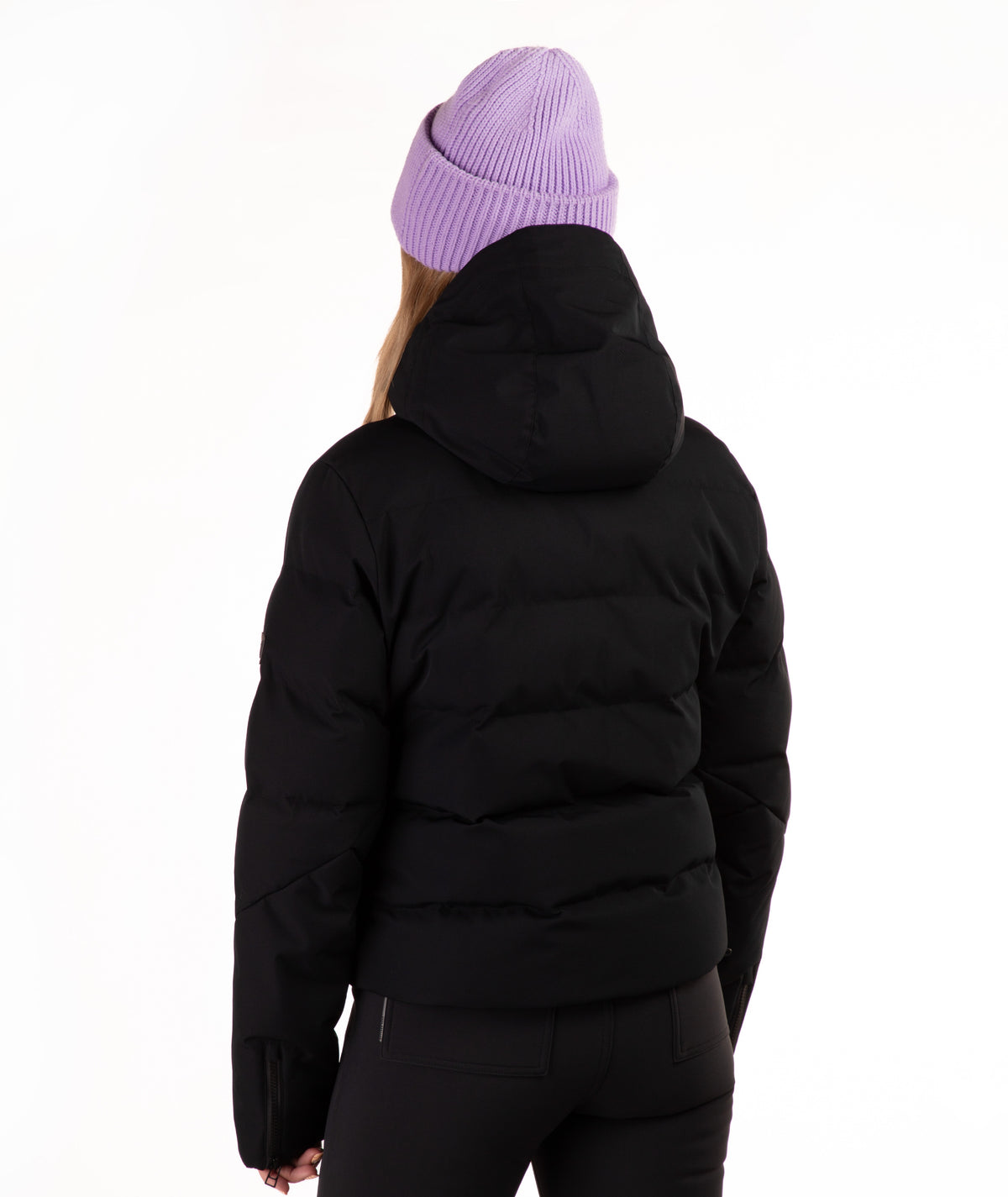A person shown from the back wearing a black hooded puffer jacket, black pants, and a light purple ribbed knit beanie against a plain white studio background.