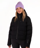 A woman wearing a black hooded puffer jacket, black pants, and a light purple knit beanie, standing against a plain white studio background.