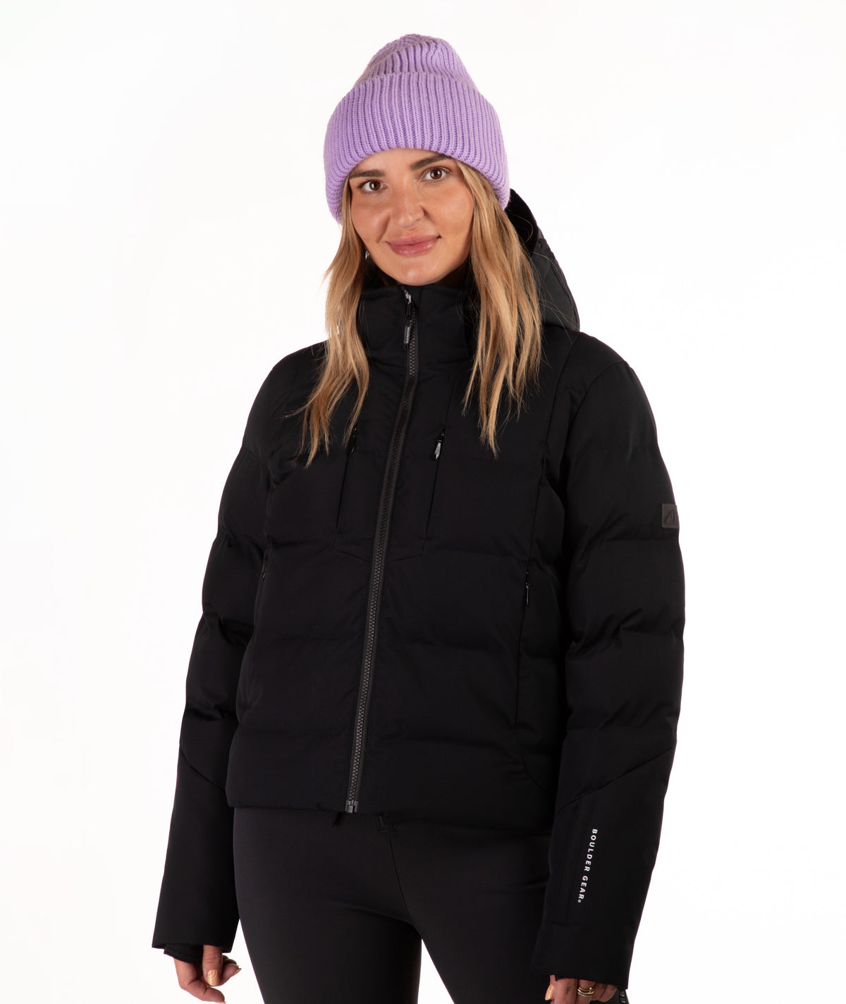 A woman wearing a black hooded puffer jacket, black pants, and a light purple knit beanie, standing against a plain white studio background.