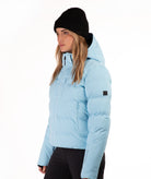 A woman wearing a light blue puffer jacket, black beanie, and black pants, shown in profile against a plain white studio background.