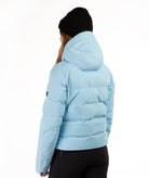 A person shown from the back wearing a light blue hooded puffer jacket, black beanie, and black pants against a plain white studio background.