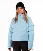 A woman wearing a light blue puffer jacket, a black beanie, and black pants, standing against a plain white studio background.