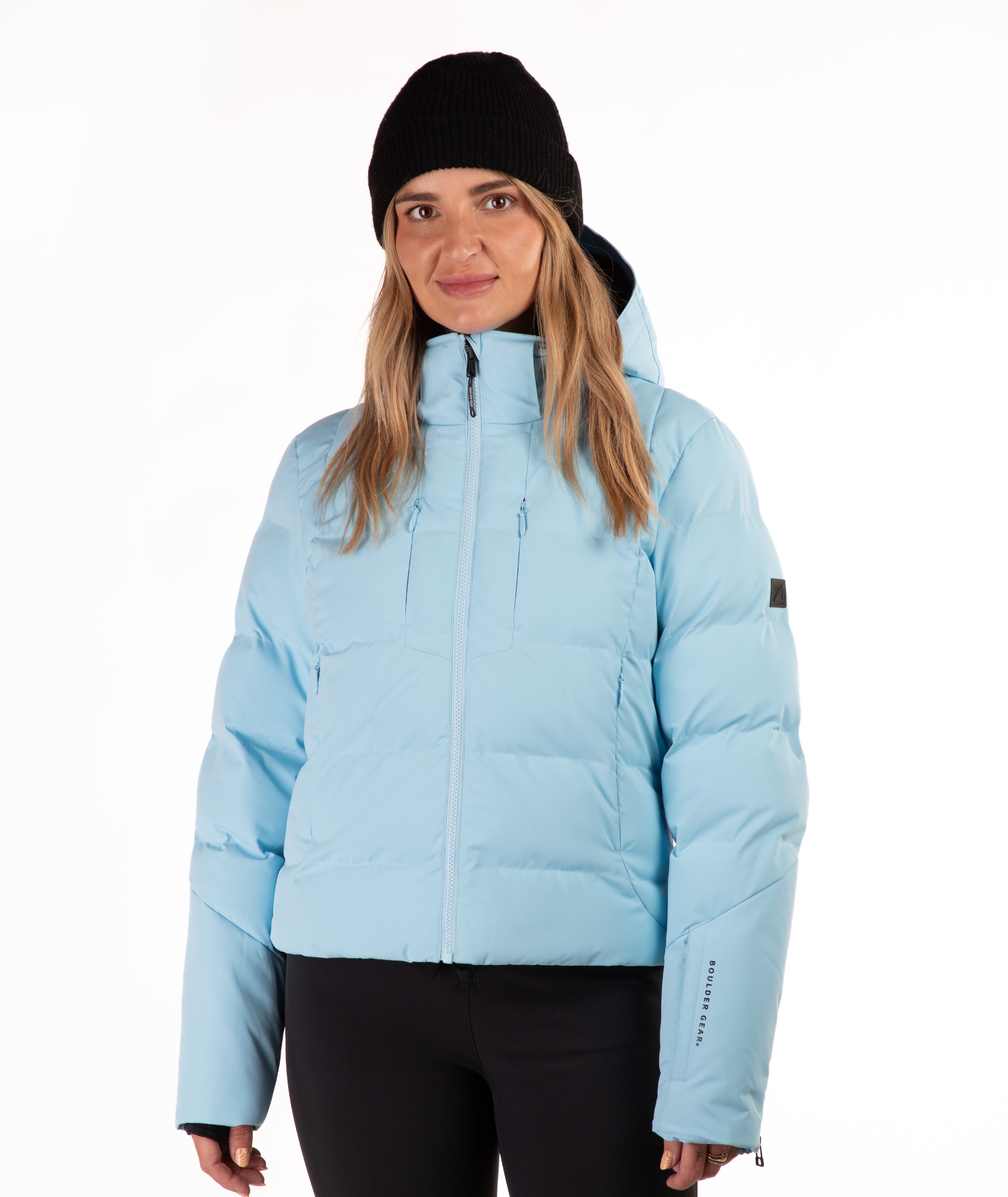 A woman wearing a light blue puffer jacket, a black beanie, and black pants, standing against a plain white studio background.