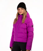 A woman wearing a bright magenta puffer jacket, a black knit beanie, and black pants, photographed against a plain white studio background.