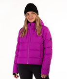A woman wearing a bright magenta puffer jacket, a black beanie, and black pants, standing against a plain white studio background.