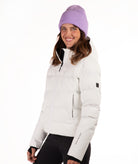 A woman wearing a off white puffer jacket and a lavender knit beanie, photographed against a clean white studio background.