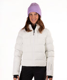 A woman wearing a off white puffer jacket, black pants, and a light purple knit beanie, standing against a plain white background.
