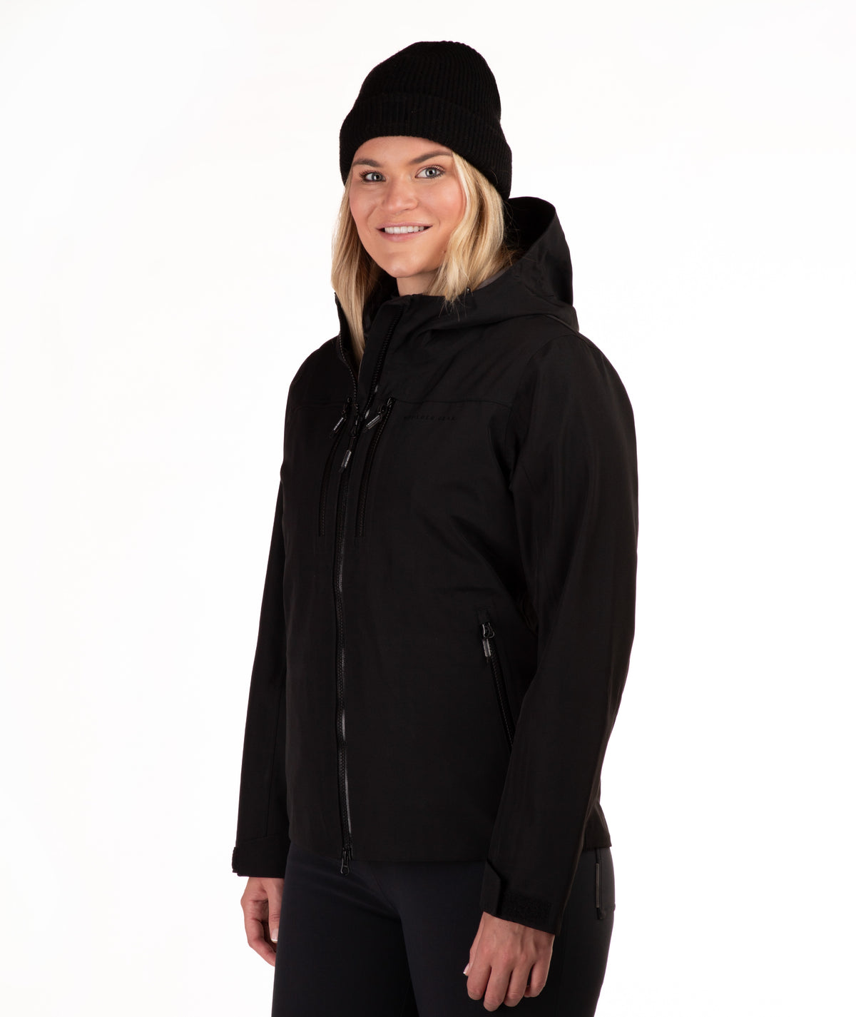 A woman wearing a black hooded jacket and a black knit beanie, photographed against a plain white studio background.