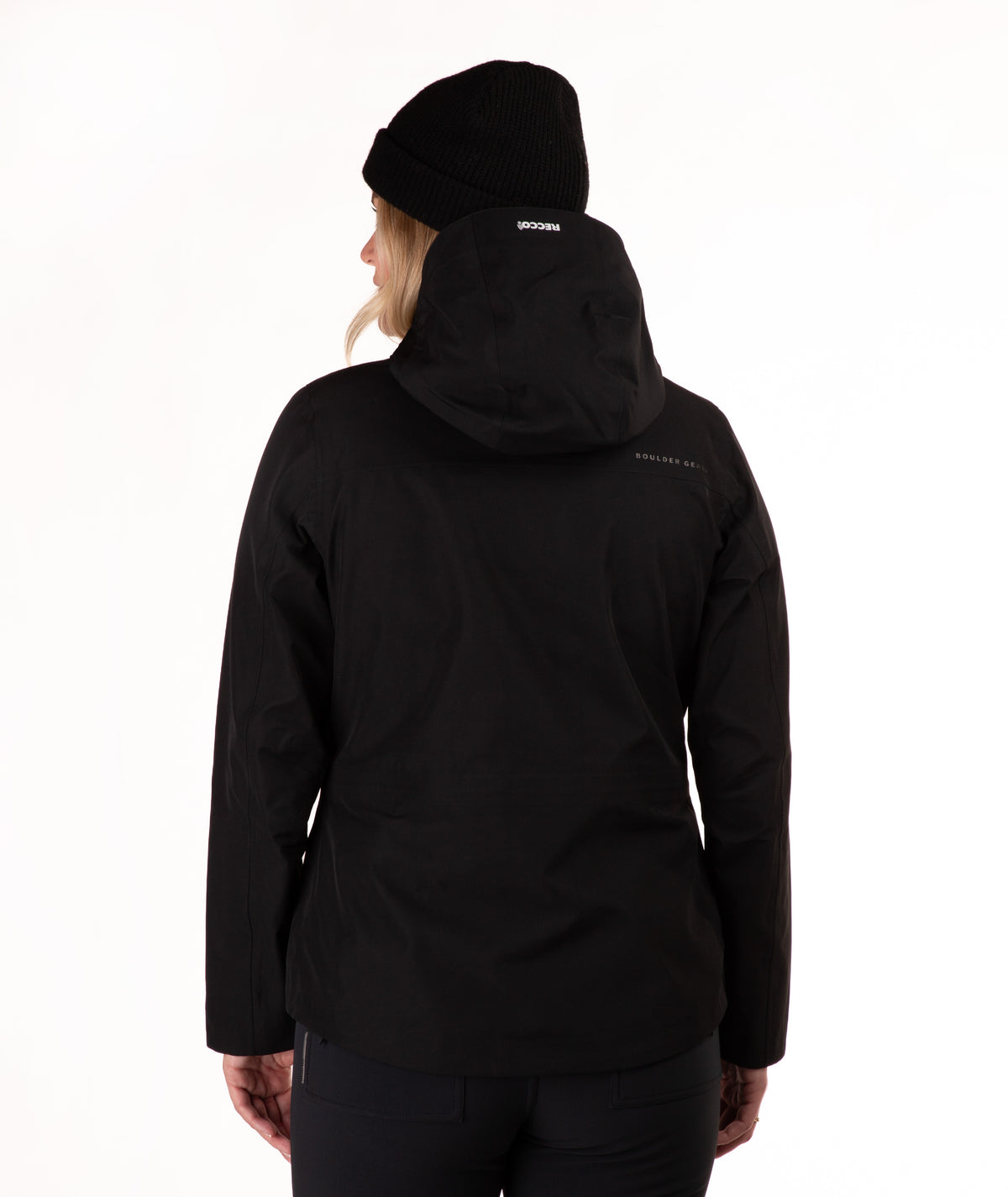 A woman viewed from the back wearing a black hooded jacket and a black beanie, photographed against a clean white studio background.