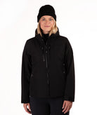 A woman wearing a black hooded zip-up jacket and a black knit beanie, standing against a plain white studio background.