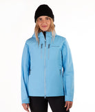 A woman wearing a light blue hooded ski jacket with a black beanie and black pants, standing against a plain white studio background.