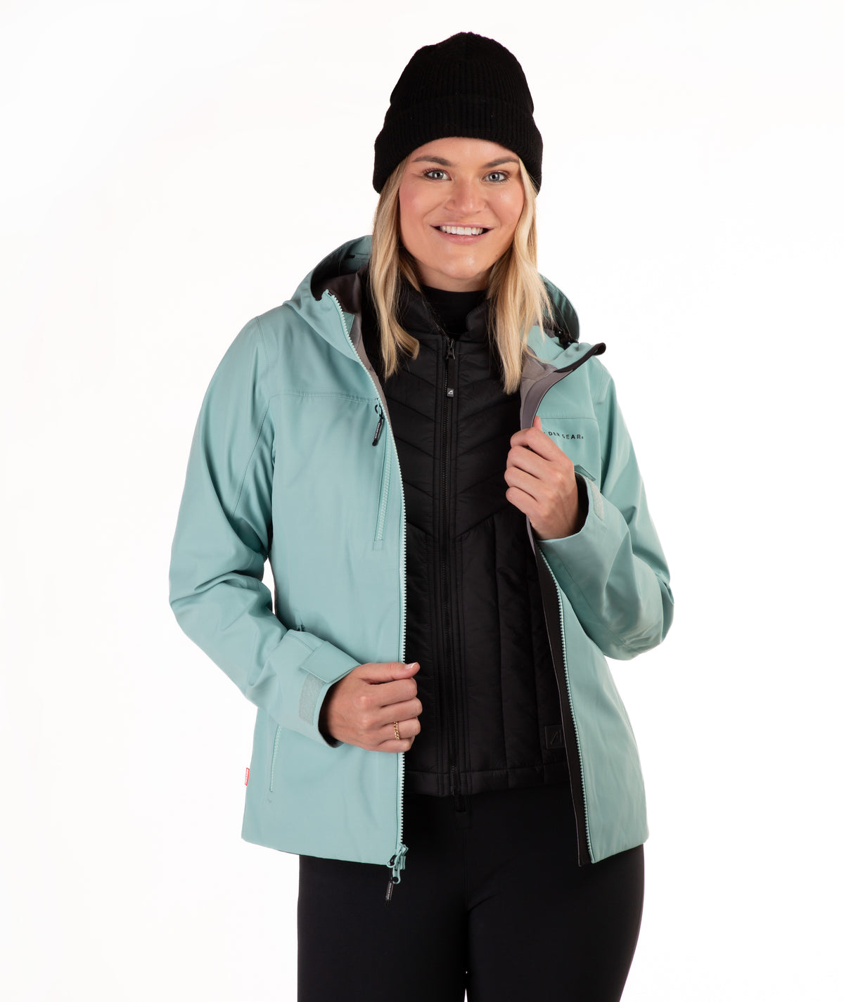 A woman wearing a light teal hooded ski jacket over a black puffer vest and black pants, paired with a black beanie, photographed against a plain white studio background.