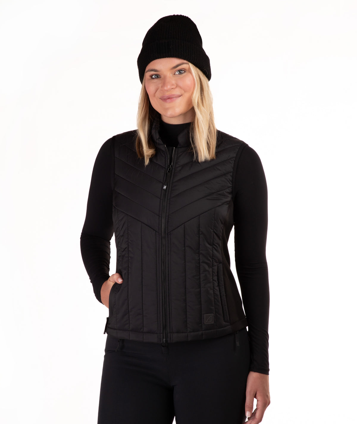 A woman wearing a black zip-front quilted vest over a black long-sleeve top and black beanie, photographed against a plain white studio background.