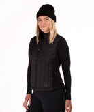 A woman wearing a black zip-front quilted vest over a black long-sleeve top and black beanie, photographed against a plain white studio background.