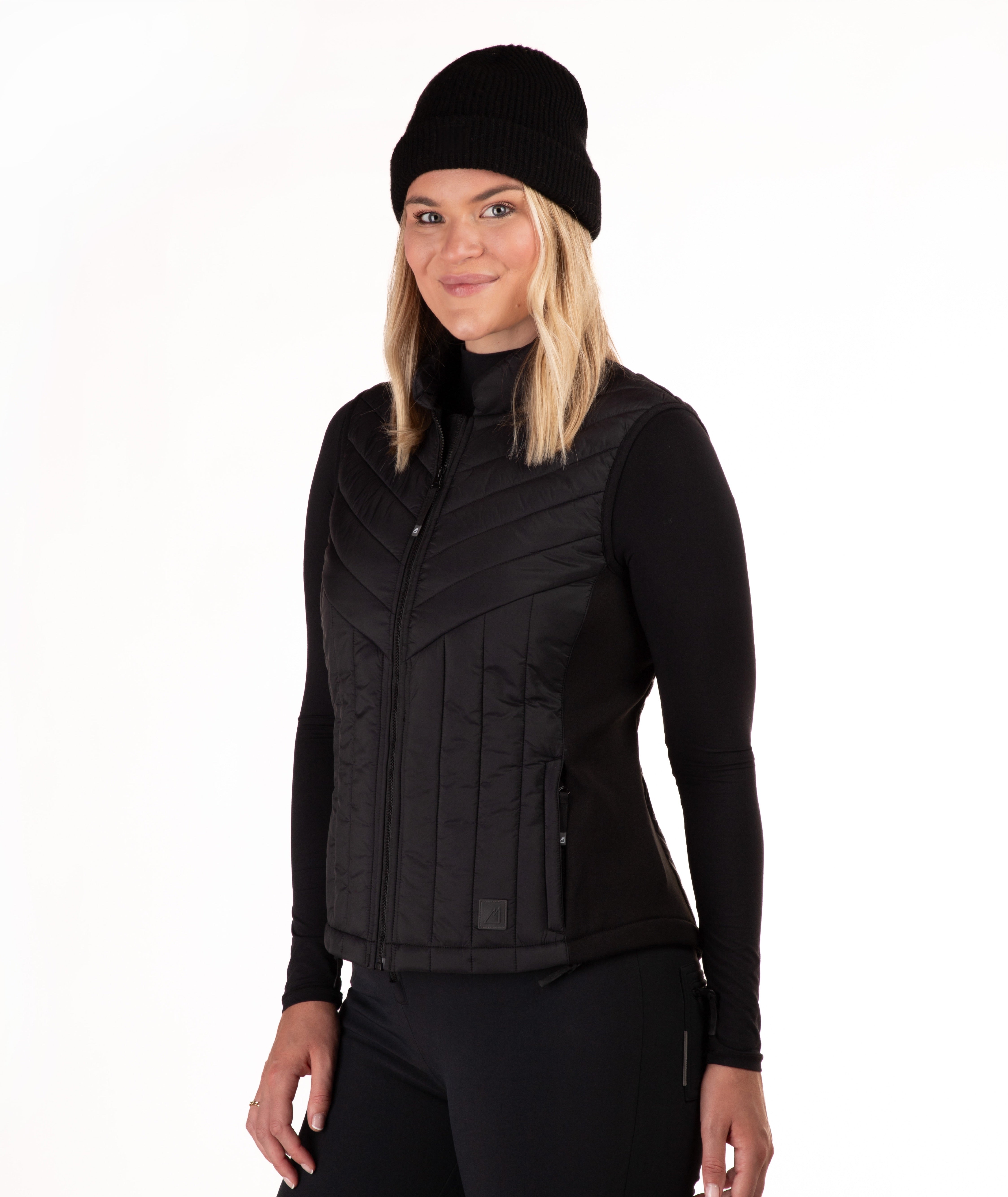 A woman wearing a black zip-front quilted vest over a black long-sleeve top and black beanie, photographed against a plain white studio background.