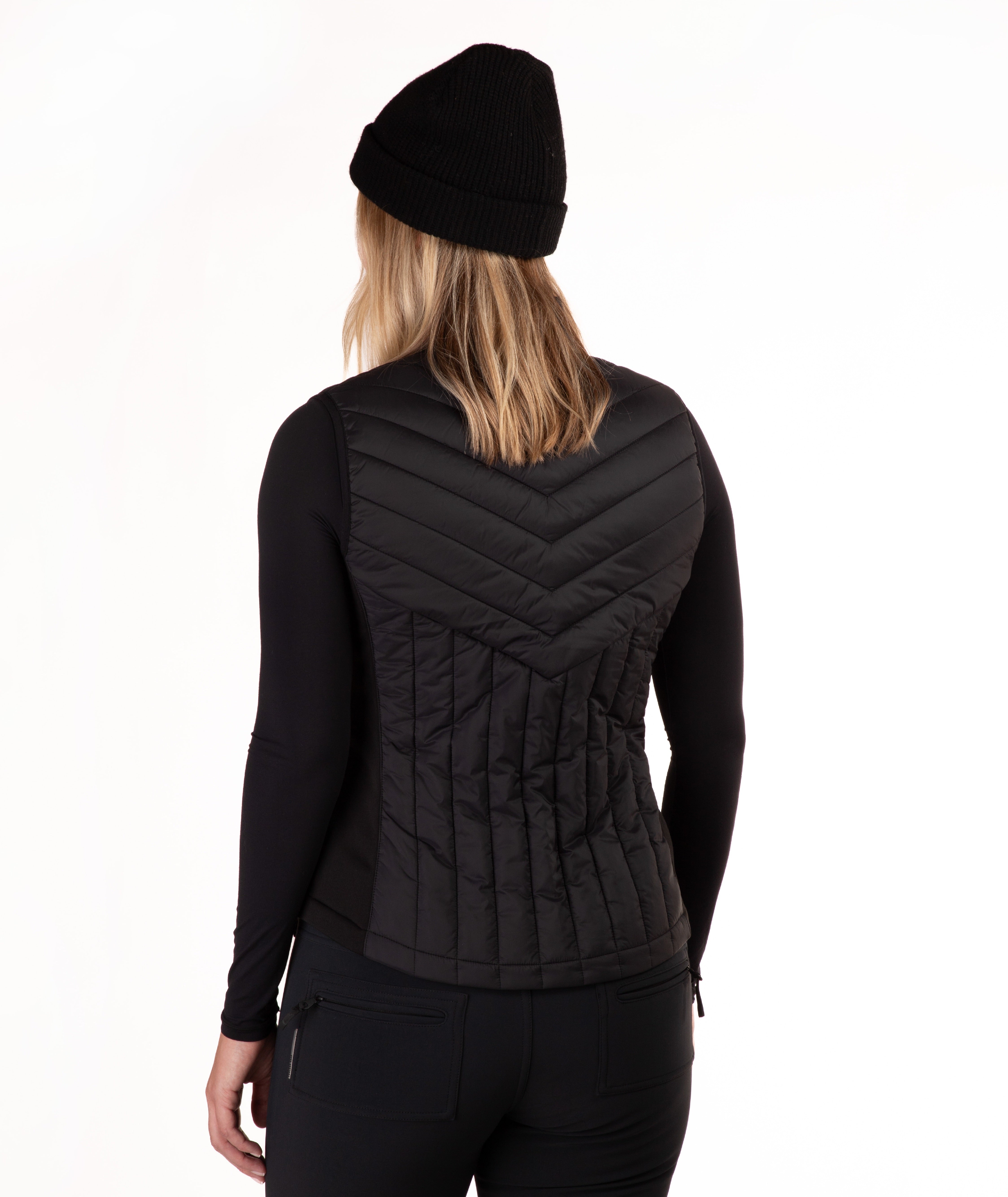 A woman wearing a black zip-front quilted vest over a black long-sleeve top and black beanie, photographed against a plain white studio background.