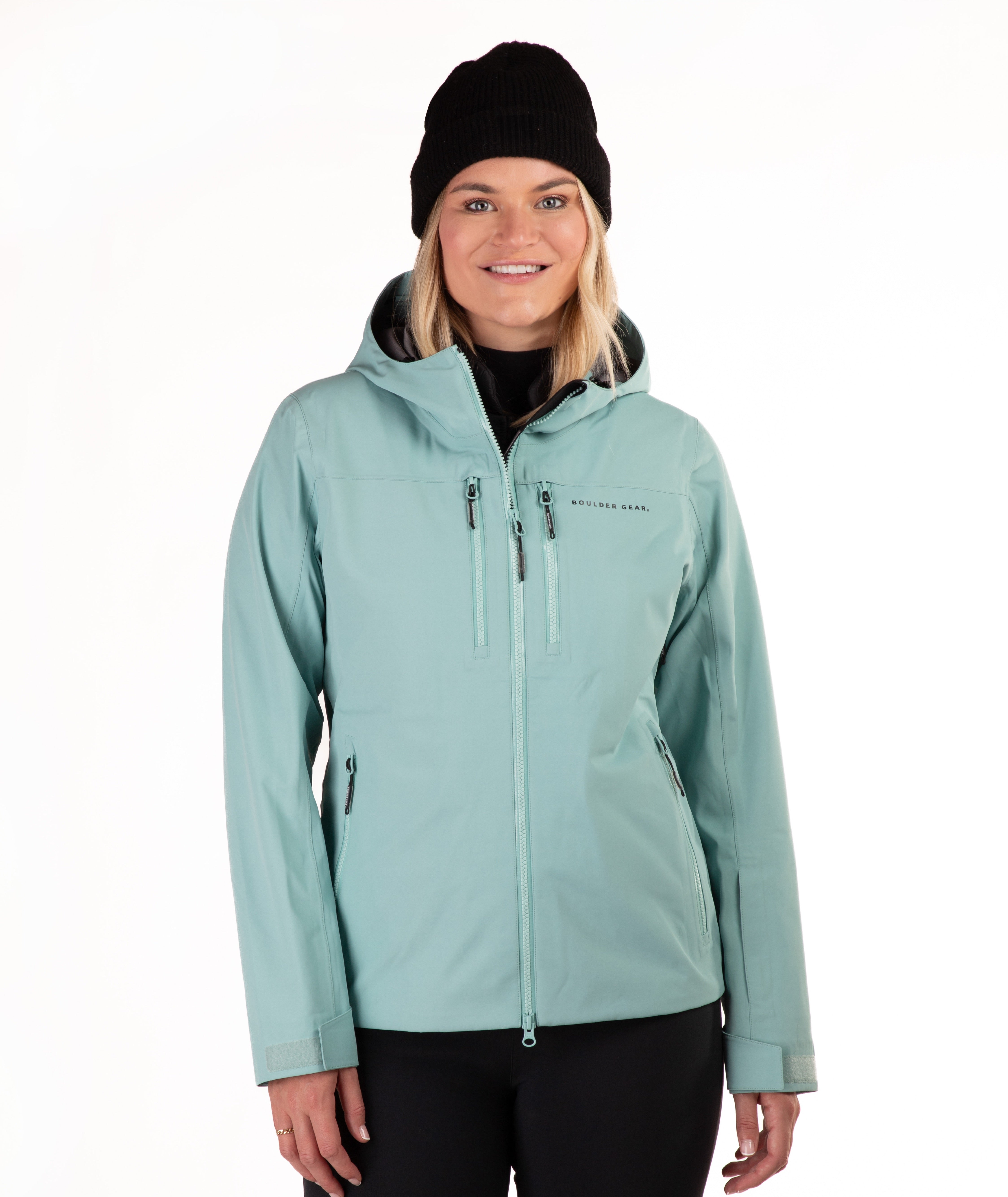 A woman wearing a light teal hooded ski jacket and black pants, paired with a black beanie, photographed against a plain white studio background.