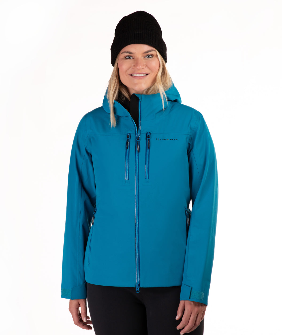 A woman wearing a bright teal hooded ski jacket, a black beanie, and black pants, standing against a plain white studio background.