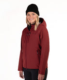 A woman wearing a burgundy hooded softshell jacket, black beanie, and black pants, standing against a plain white studio background.