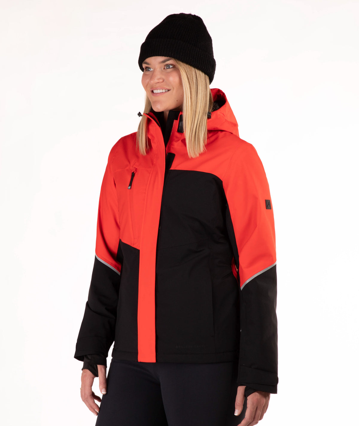 A woman wearing a bright red and black color-blocked ski jacket, a black beanie, and black pants, photographed against a clean white studio background.