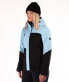 A woman wearing a light blue and black color-blocked ski jacket, a black beanie, and black pants, photographed against a clean white studio background.