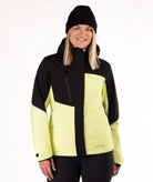 A woman wearing a black and light yellow-green color block ski jacket, a black beanie, and black pants, photographed against a plain white studio background.