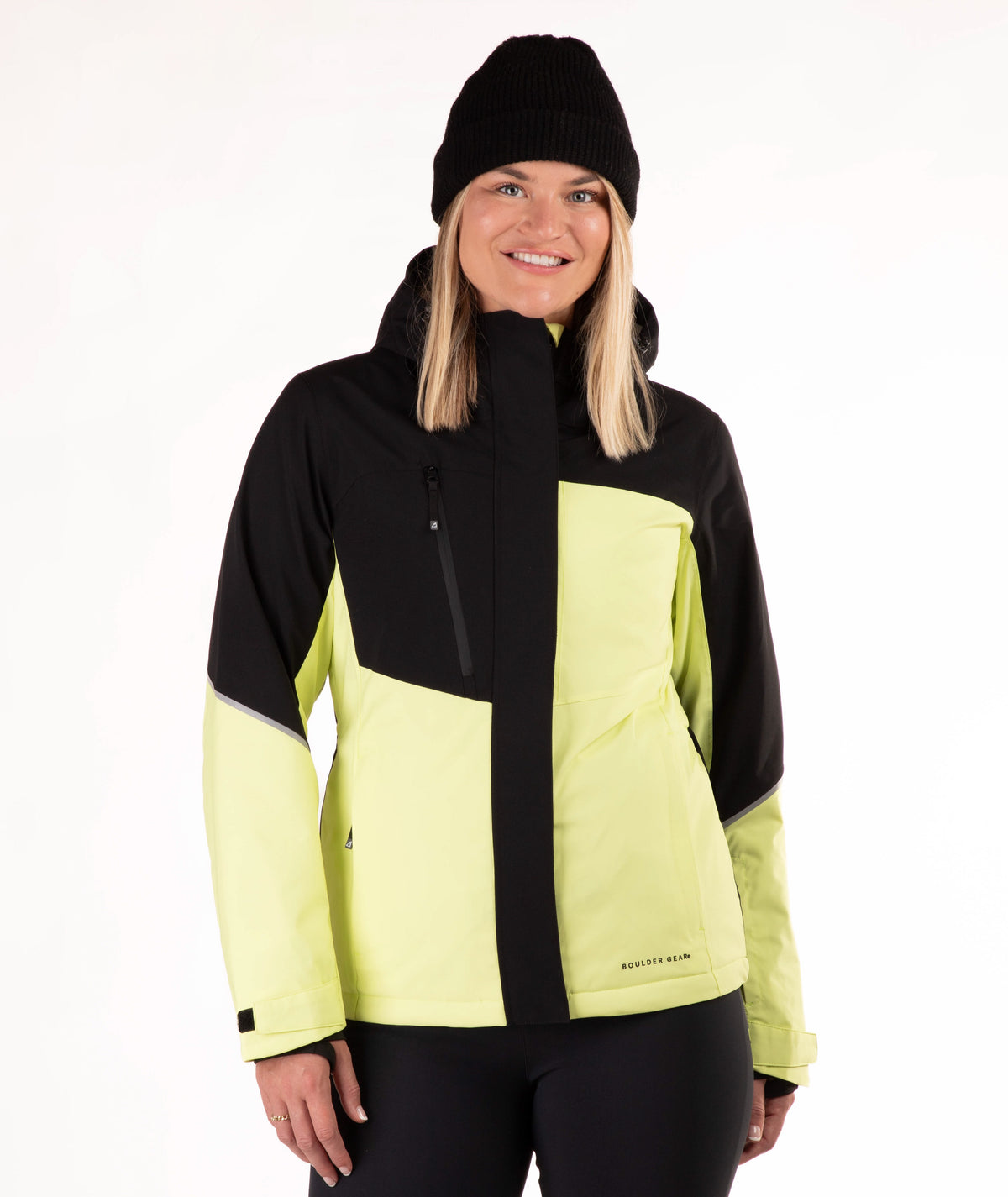 A woman wearing a black and light yellow-green color block ski jacket, a black beanie, and black pants, photographed against a plain white studio background.
