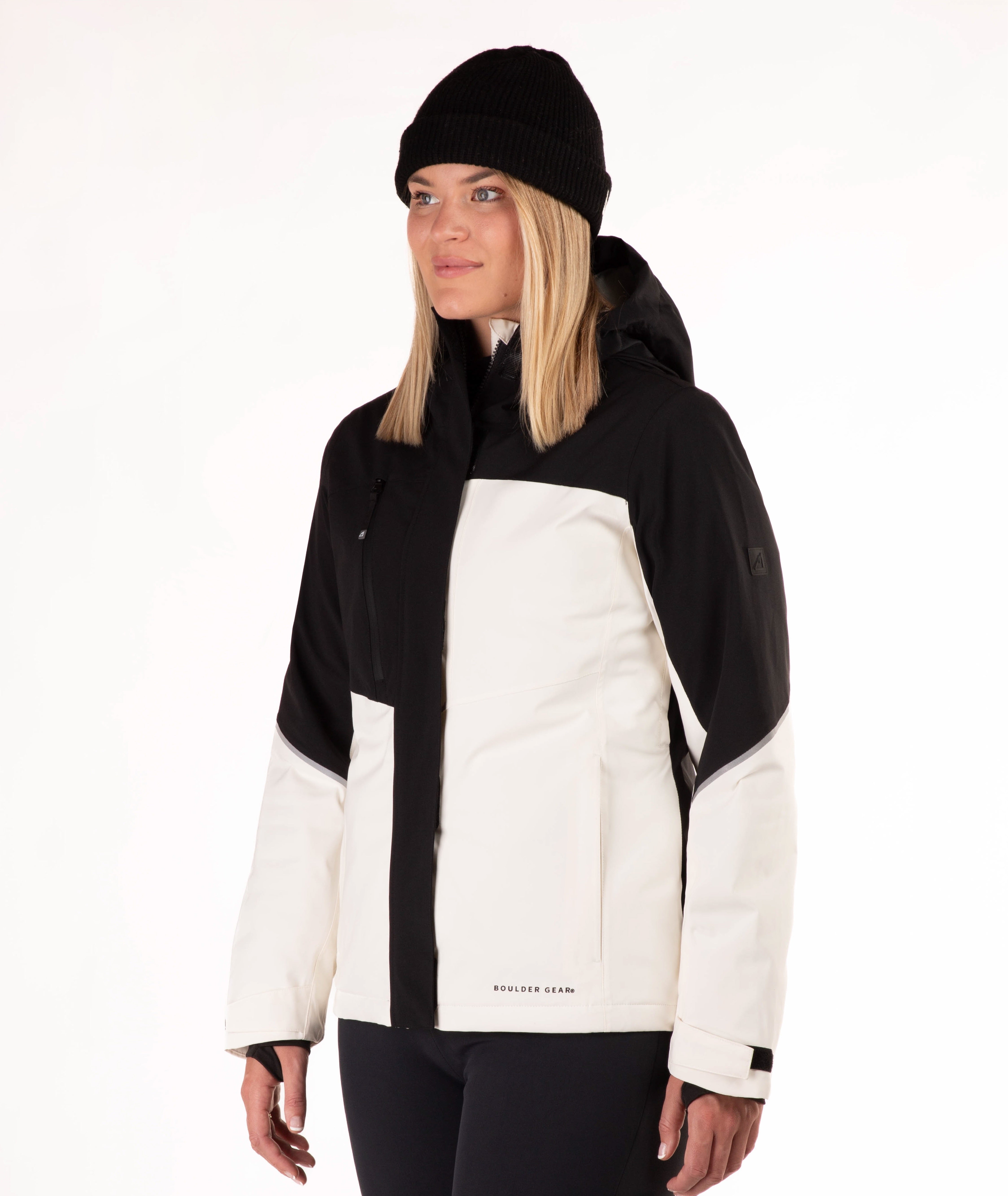 A woman wearing a black and off-white color-blocked ski jacket, a black beanie, and black pants, photographed against a clean white studio background.