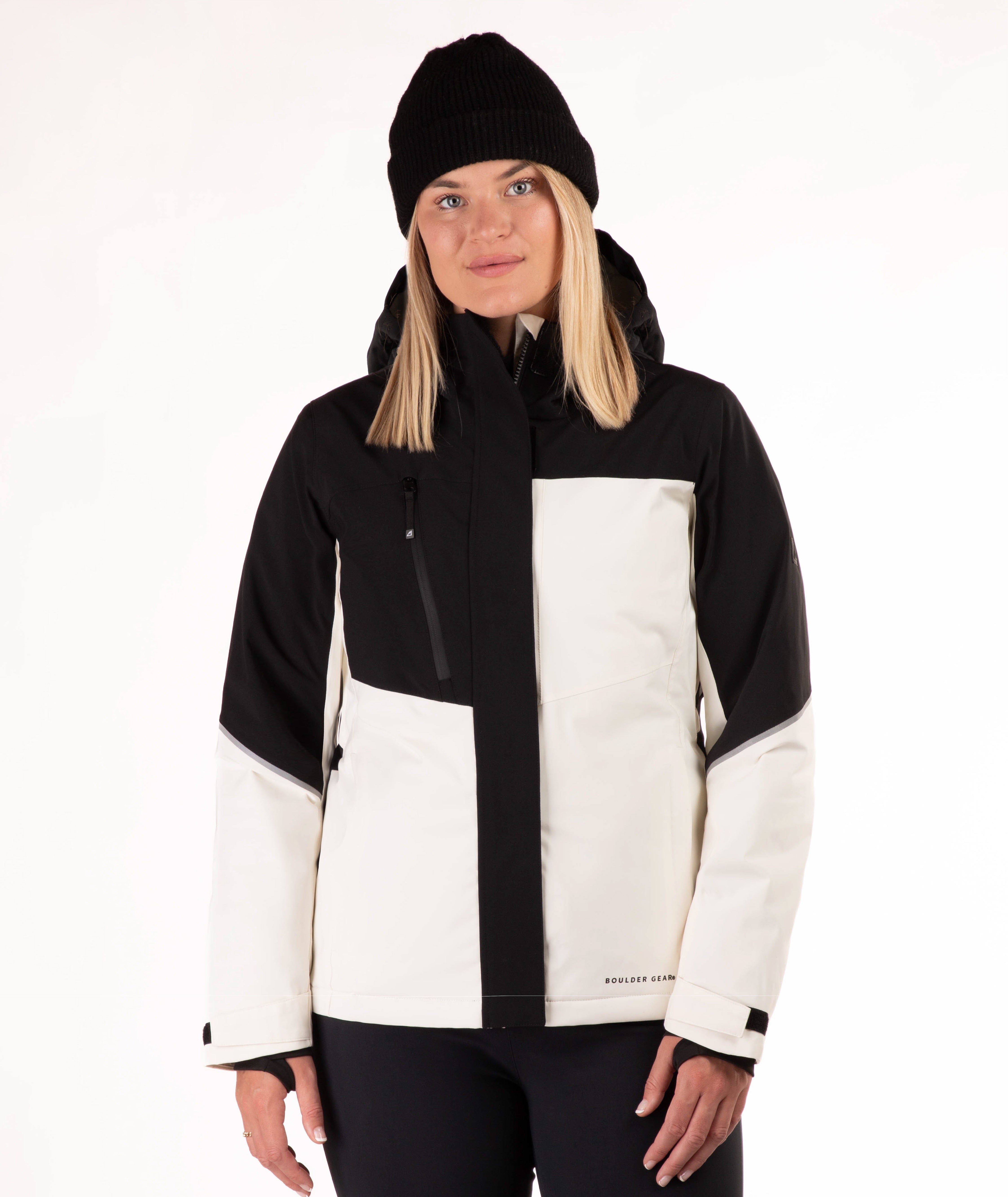 A woman wearing a black and off-white color-blocked ski jacket, a black beanie, and black pants, photographed against a plain white studio background.