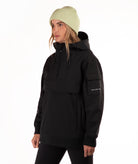 A woman wearing a black hooded pullover jacket with a high collar, drawstrings, and a horizontal chest pocket, paired with a light green ribbed knit beanie against a plain white background.