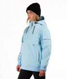 A woman wearing a light blue hooded pullover jacket with black drawstrings, a black beanie, and black pants, photographed against a plain white studio background.