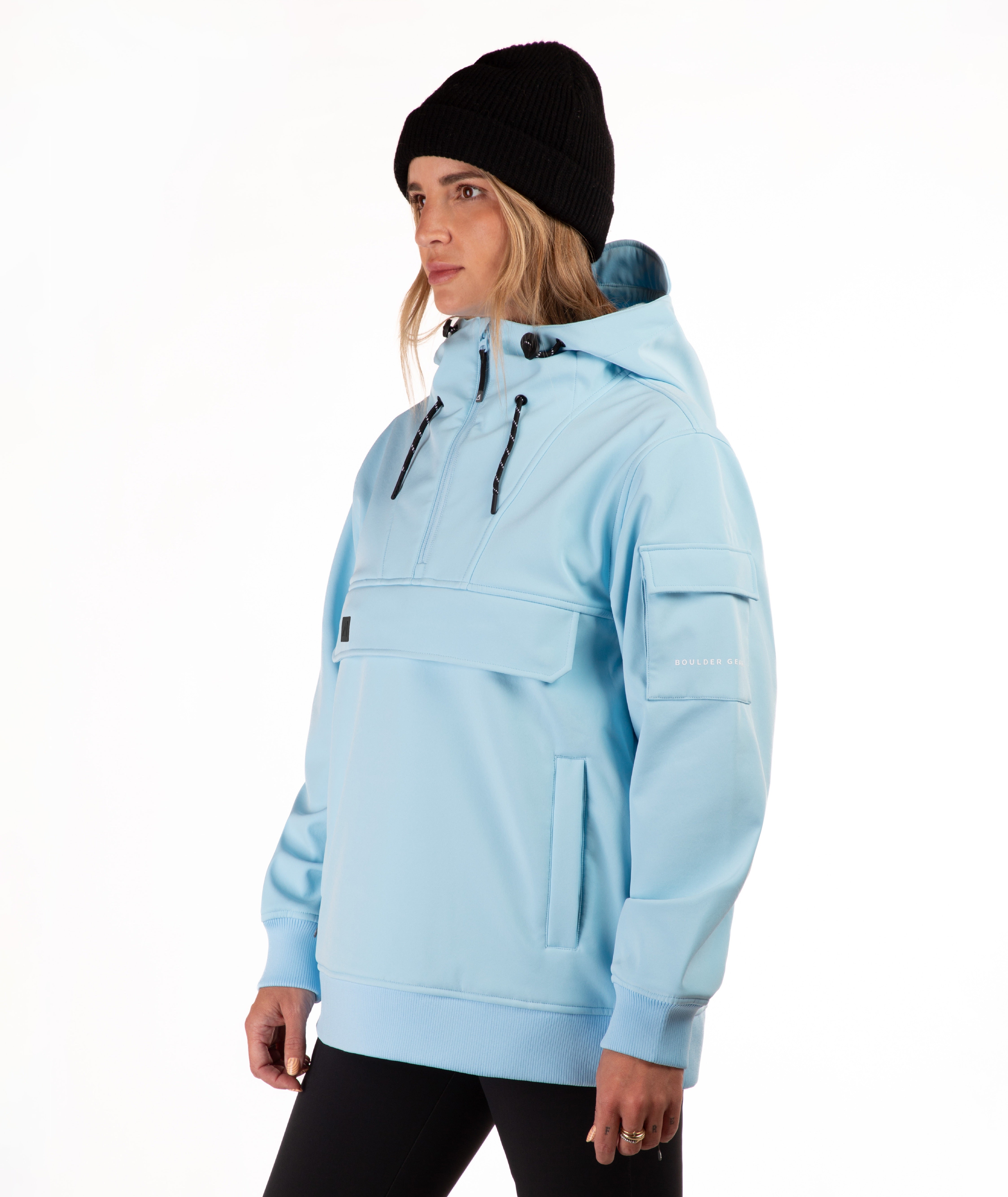 A woman wearing a light blue hooded pullover jacket with black drawstrings, a black beanie, and black pants, photographed against a plain white studio background.