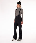 Woman wearing black ski bib overalls with a gray top on a white background