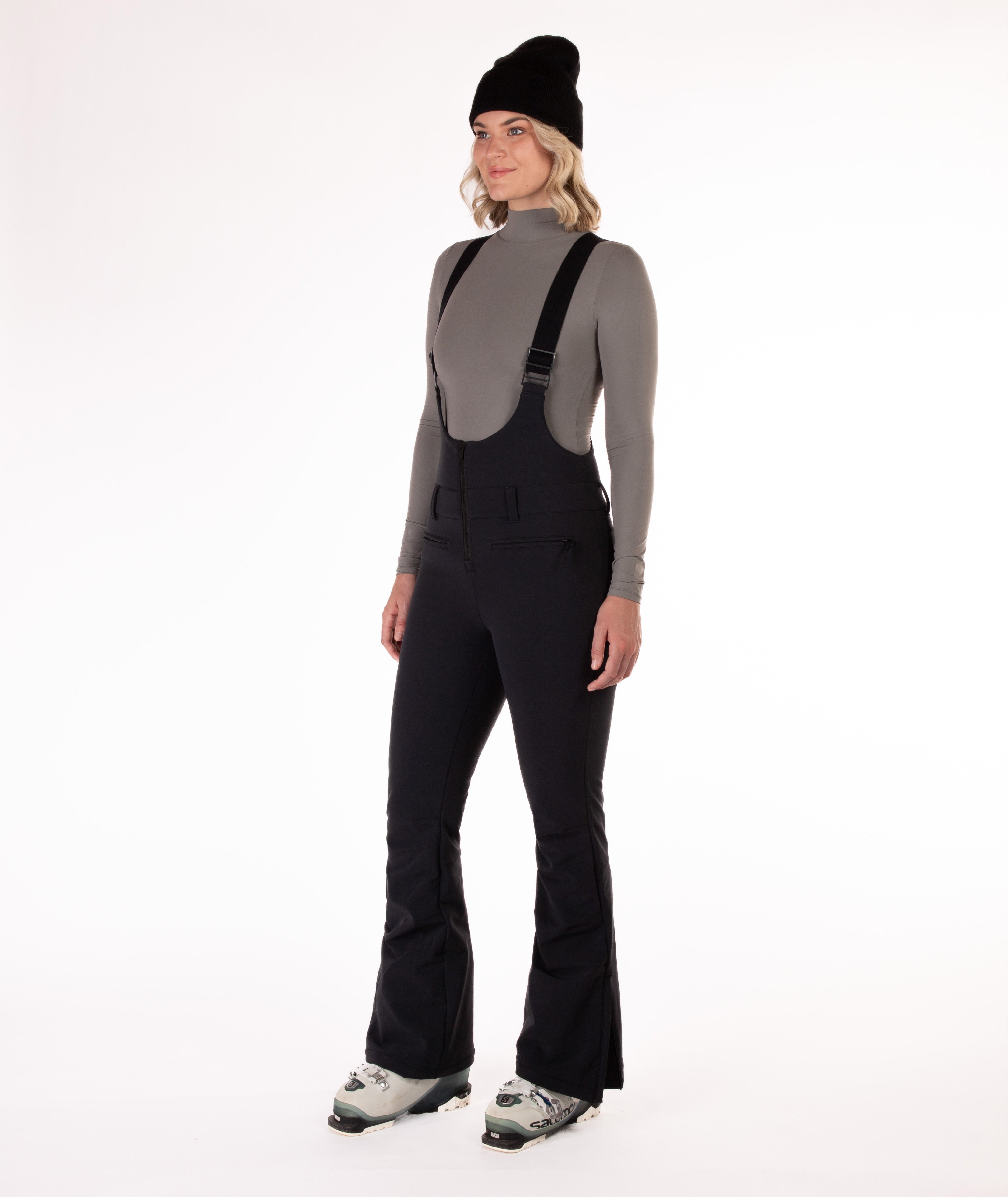 Woman wearing black ski bib overalls with a gray top on a white background