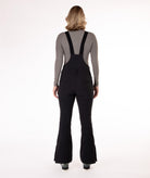 back of a Woman wearing black ski bib overalls with a gray top on a white background