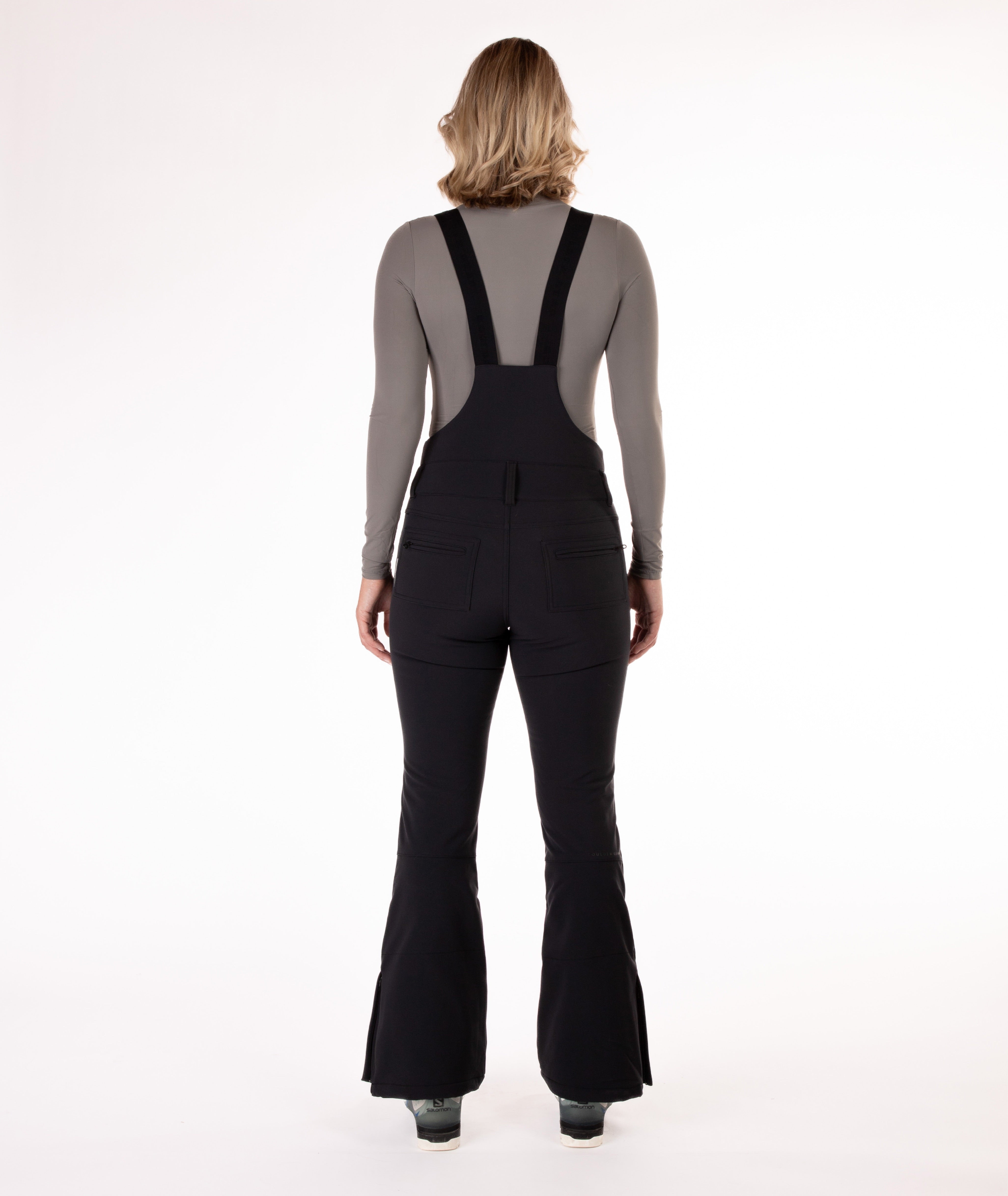 back of a Woman wearing black ski bib overalls with a gray top on a white background