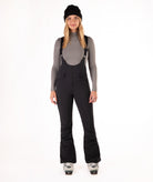 Woman wearing black ski bib overalls with a gray top on a white background