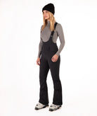 Woman wearing black ski bib overalls with a gray top on a white background